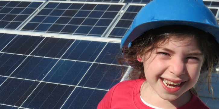 solar for schools and colleges. An image of a little girl looking happy whilst stood infront of solar panels, learning about renewable energy.