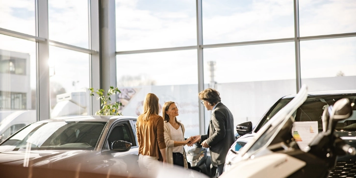 Solar panels for Car Dealerships. An image of a businessman with short brown hair and glasses is giving the keys to a female customer with long brown hair, both smiling at each other, woman with blonde hair standing in the front, indoors at a car dealership. 